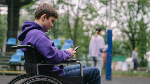 een man zit in een rolstoel met zijn mobiel in zijn hand. Hij bevindt zich in een parkachtige omgeving met veel bomen.