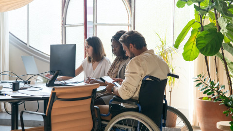 Aan een bureau zitten drie personen waarvan 1 man in een rolstoel. Ze zijn aan het werk op een computer en tablet.