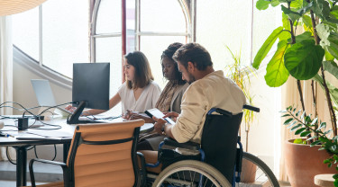 Aan een bureau zitten drie personen waarvan 1 man in een rolstoel. Ze zijn aan het werk op een computer en tablet.