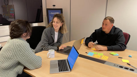 Drie mensen zitten met laptops aan tafel in een kantoorruimte. Ze zijn in gesprek en lachen. Op tafel liggen posts-its.