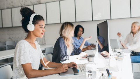 Een bureau waaraan een tester software aan het testen is achter de computer en op de achtergrond zijn drie collega's aan het overleggen.