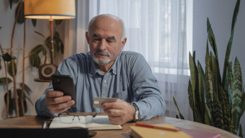 Een man (senior) zit aan tafel met zijn mobiele telefoon en bankpas in zijn hand, te bedenken hoe hij hier nu verder mee moet.