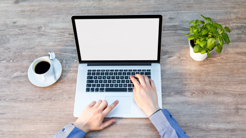 Op een tafel staat een laptop, kop koffie en plantje. De laptop wordt bedient door twee handen.