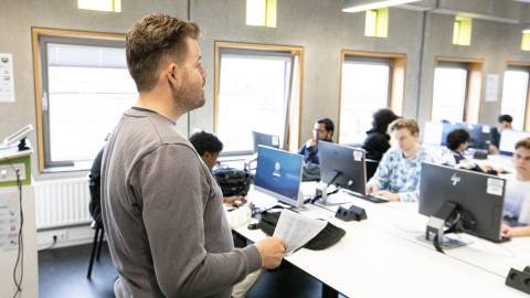 Docent staat voor de klas, jongens zitten achter computerscherm