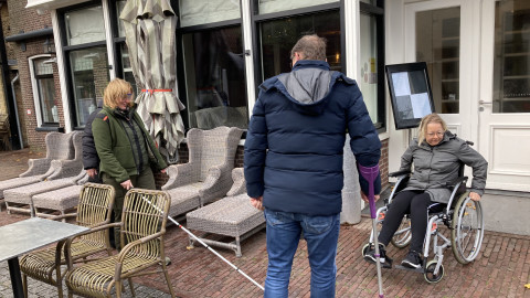 Een foto in de winkelstraat op Vlieland waar een persoon met witte stok loopt, een persoon in een rolstoel rondrijdt en een begeleider te zien is