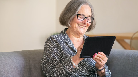 Oudere vrouw met bril zit op bank met tablet in haar hand