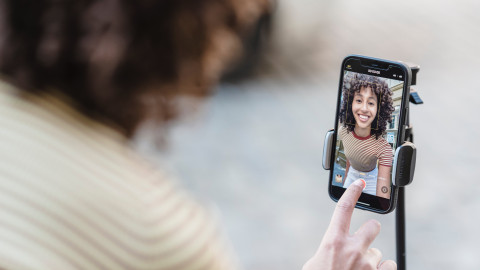 Je ziet een mobiel op een selfie stick waarop een vrouw een video van zichzelf opneemt. Haar vinger drukt op het afspeelknopje om de video op te nemen.