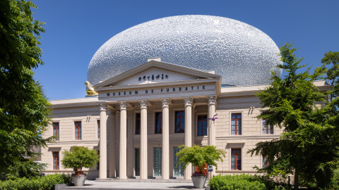 het statige gebouw van museum de fundatie in zwolle met zes zuilen voor de ingang en bovenop het gebouw staat een witkleurige bol dat een mooi contrast vormt met het zandkleurige statige gebouw.