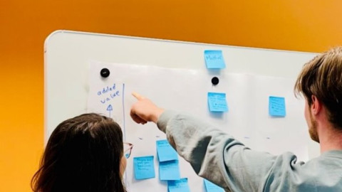 Een whiteboard hangt tegen een gele muur. Er staan twee personen voor het whiteboard met hun rug naar de camera toe. De ene persoon wijst naar een groot papier met blauwe post-its die op het whiteboard hangt. De andere persoon kijkt mee.