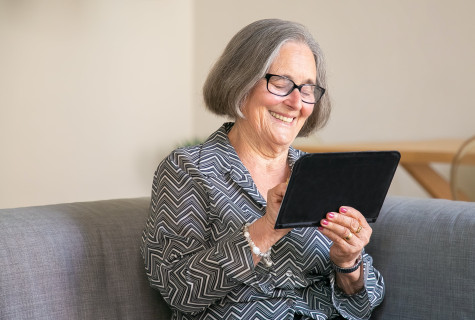 Oudere vrouw met bril zit op bank met tablet in haar hand