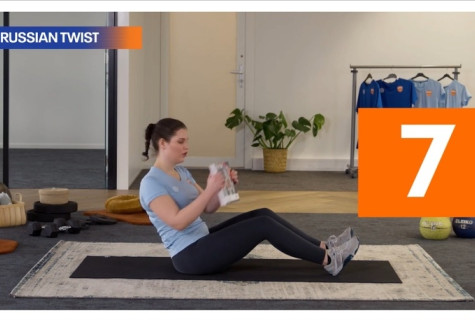 Een dame in zwarte broek en lichtblauw shirt en sportschoenen zit op de grond met haar knieen licht gebogen en een rechte rug. Ze houdt een boek in haar handen en doet de sportoefening russian twist.