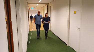 Man met geleidestok en vrouw zonder stok lopen door een gang in een kantoorpand.