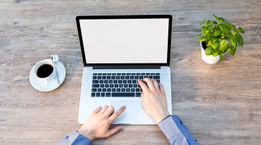 Laptop van bovenaf gezien. Twee handen hangen boven het toetsenbord en de mousepad. De laptop staat op een tafel. Naast de laptop staan een kopje koffie en een plantje.