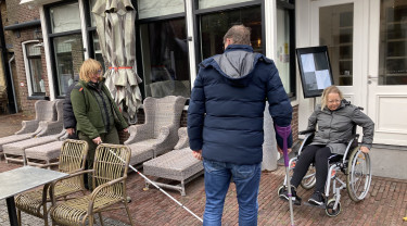 Een foto in de winkelstraat op Vlieland waar een persoon met witte stok loopt, een persoon in een rolstoel rondrijdt en een begeleider te zien is