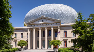 het statige gebouw van museum de fundatie in zwolle met zes zuilen voor de ingang en bovenop het gebouw staat een witkleurige bol dat een mooi contrast vormt met het zandkleurige statige gebouw.