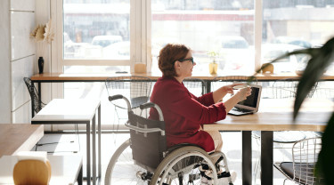 een dame in een rolstoel zit aan een tafel in een open ruimte op haar laptop