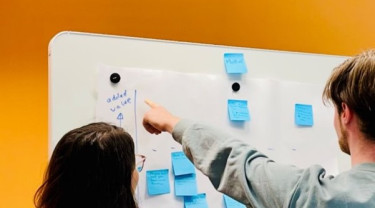 Een whiteboard hangt tegen een gele muur. Er staan twee personen voor het whiteboard met hun rug naar de camera toe. De ene persoon wijst naar een groot papier met blauwe post-its die op het whiteboard hangt. De andere persoon kijkt mee.