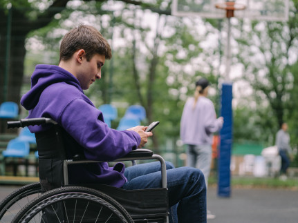 een man zit in een rolstoel met zijn mobiel in zijn hand. Hij bevindt zich in een parkachtige omgeving met veel bomen.