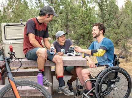 Een man zit op tafel, een vrouw zit aan tafel, een man zit in rolstoel aan tafel, buiten in een park, en ze drinken een biertje