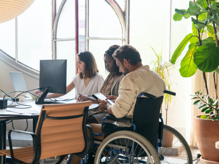 drie collega's aan het werk bij een bureau waarop twee computers staan en een van de collega's aan tafel zit in een rolstoel en bedient een ipad