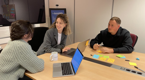 Drie mensen zitten met laptops aan tafel in een kantoorruimte. Ze zijn in gesprek en lachen. Op tafel liggen posts-its.