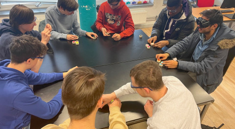 aan tafel zitten 8 studenten met een bril voor troebel zicht een voelspelletje te doen