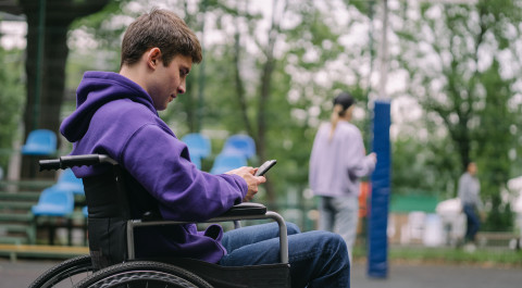 een man zit in een rolstoel met zijn mobiel in zijn hand. Hij bevindt zich in een parkachtige omgeving met veel bomen.