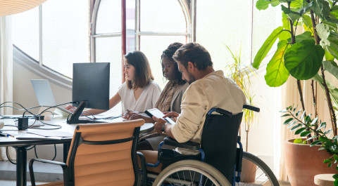 Aan een bureau zitten drie personen waarvan 1 man in een rolstoel. Ze zijn aan het werk op een computer en tablet.