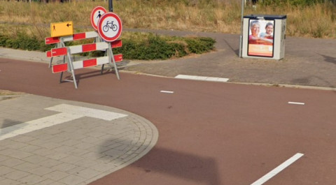 Een onveilige en onoverzichtelijke verkeerssituatie voor voetgangers met obstakels zoals een verkeersbord op het fietspad en een elektriciteitskast op de stoep aan het einde van de geleidelijnen.