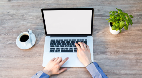 Op een tafel staat een laptop, kop koffie en plantje. De laptop wordt bedient door twee handen.