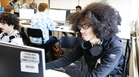 Een jongeman zit in een leslokaal achter een computer waarop hij aan het werk is. Om hem heen zitten andere studenten achter de computer.
