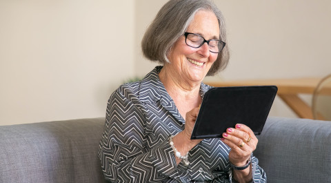 Oudere vrouw met bril zit op bank met tablet in haar hand