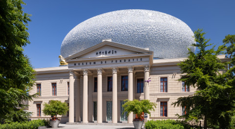 het statige gebouw van museum de fundatie in zwolle met zes zuilen voor de ingang en bovenop het gebouw staat een witkleurige bol dat een mooi contrast vormt met het zandkleurige statige gebouw.
