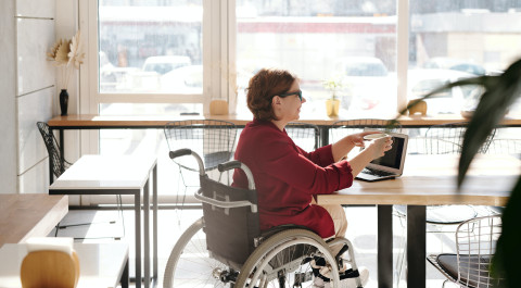 een dame in een rolstoel zit aan een tafel in een open ruimte op haar laptop