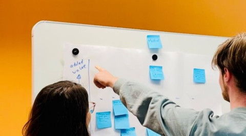 Een whiteboard hangt tegen een gele muur. Er staan twee personen voor het whiteboard met hun rug naar de camera toe. De ene persoon wijst naar een groot papier met blauwe post-its die op het whiteboard hangt. De andere persoon kijkt mee.