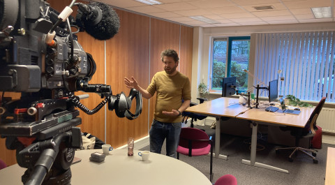 in een kantoor staat Martijn voor een videocamera op een statief met op de achtergrond twee bureaus in beeld.